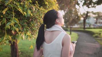 Disparo por detrás de una joven asiática con ropa de ejercicio que usa auriculares con un teléfono inteligente mientras se refresca en el parque durante la cálida hora del atardecer, caminata relajante limpiándose el sudor de la cara después del ejercicio video