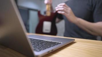 nahaufnahme eines verschwommenen männlichen schwarzen hemdkörpers mit computer-laptop mit haltender e-gitarre, während er auf einem holzstuhl sitzt, kostenlose online-akkorde im internet, ein online-entertainer, live-streaming