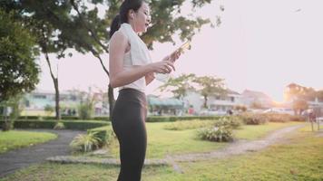 asiatisk ung kvinna bär sportkläder med hjälp av smartphone medan de tränar i utomhusparken, vid solnedgången koppla av singel kvinna livsstil, trådlös digital teknik bärbar gadget mobilapplikation användare video