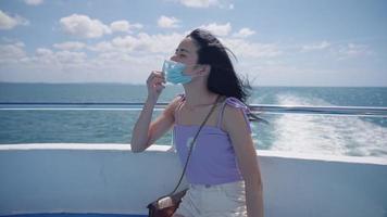 A young attractive black hair female slowly removing a medical mask and breathing deeply an ocean breeze, enjoying idyllic natural resources view, a summer cruise trip, woman relaxing on boat deck