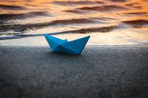 Paper blue ship is thrown on sandy shore of lake at sunset.