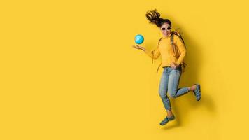 Woman traveler with suitcase, holding ball globe in the hand on Yellow background. Portrait of smiling happy girl with space copy the text. Travel backpack photo
