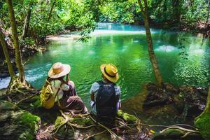 Travelers couples with backpacks sitting and relax on the rocks. travel nature in greens jungle and enjoying view in waterfall. Tourism, hiking, nature study. Couples traveling taking pictures