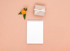 un cuaderno blanco vacío, una mandarina y un pequeño regalo sobre un fondo de melocotón. plantilla con espacio para texto, concepto de planificación o maqueta. foto