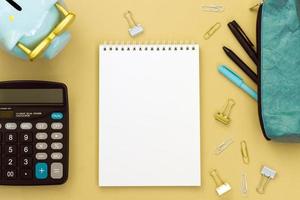 The concept of planning. Top view of a Desk with a pencil case, an empty Notepad, a piggy Bank and a calculator on a yellow background. Flat lay of items with space for text.