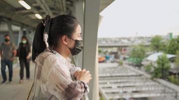 una joven asiática usa una máscara negra apoyándose en cruzar el borde del puente pensando en mirar hacia la carretera, riesgo de infección en lugares públicos, precaución al salir, tiro al aire libre con gente caminando en el fondo video