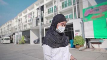 junge asiatin trägt schwarzen hijab und schützende gesichtsmaske, benutzt handy beim gehen mit geparktem auto im städtischen hintergrund, netzwerkverbindung, covid-19-pandemie neue normalität, soziale distanzierung