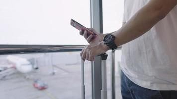 giovane maschio in piedi all'interno della sala partenze del terminal dell'aeroporto utilizzando smartphone, volo aereo viaggio vacanza con bagaglio a mano, prenotazione prenotazione biglietti, spostamento a bordo dell'immigrazione video