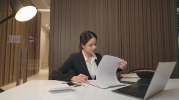 Asian success entrepreneur woman inspecting company report sheet with unhappy expression. Office management woman reading unhappy with paper documents while having online conference during night time