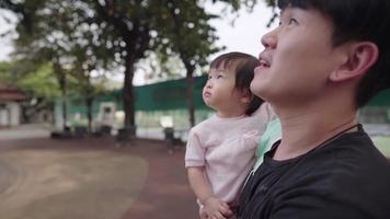 jovem pai asiático segurando sua filhinha olhando para o céu dentro do parque público em pé debaixo de árvores, ligação de cuidados infantis, inocência de crianças, seguro de vida de cena feliz de pai e filha