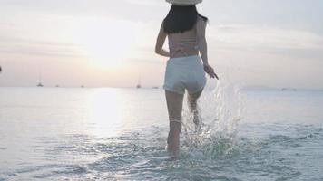 Asian young woman running into the ocean on the tropical island beach, sea water splashing in slow motion on sunset hour. Summer holiday travel destination paradise on earth fun , escape from all