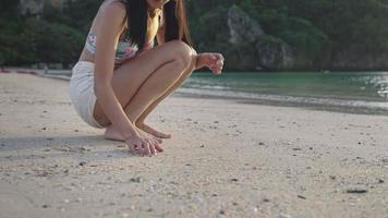 mujer asiática atractiva en bikini sentada en cuclillas jugando en arena empapada contra la luz del sol hora dorada, destino de viaje paradisíaco de isla tropical, actividad de ocio en la playa, vacaciones de verano, naturaleza asiática