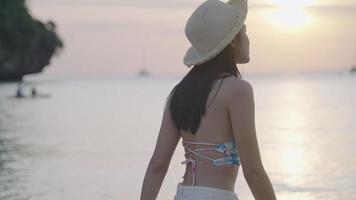 jovem asiática usa chapéu desfrutar de vibração de praia sorrindo para a câmera, virando o corpo olhando para o oceano na hora do pôr do sol. sentindo relaxamento alívio pulando em volta do corpo, estilo de vida de liberdade,