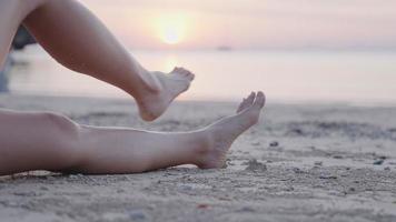 Cierra las piernas femeninas yaciendo aisladas en la playa tropical de arena de la tarde, un turista relajante viendo la puesta de sol solo en la costa, una mujer asiática adulta alegre y descalza sentada sola en la arena