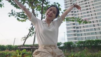 Young pretty asian woman showing happy life in city, positive energy, happy joyful girl expressing overwhelming happiness, looking at camera sitting against modern high rise condo green garden park video
