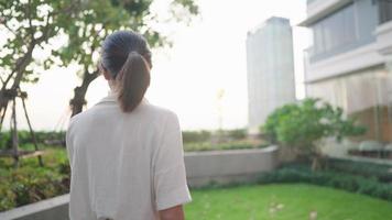 una mujer casual asiática tranquila caminando sola con un libro de lectura contra la luz del sol de la tarde en el parque del cielo del condominio, un entorno verde y un edificio moderno de gran altura en el fondo, la gente y el hobby del hogar video