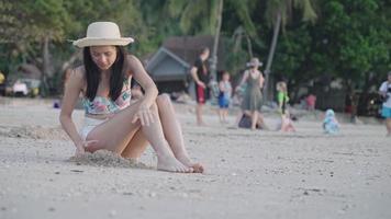 una mujer asiática delgada y juguetona cavando en una superficie de arena mientras se sienta en una playa famosa con gente contenta detrás, refrescándose con un ambiente verde, viajando en verano en la isla, relajándose el fin de semana