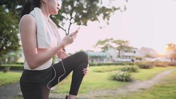 Aziatische jonge vrouw die smartphone gebruikt die muziek kiest en zich klaarmaakt om op te warmen voordat ze in het openluchtpark rent tijdens zonsondergang, ontspannende training, nieuw normaal modern leven, quality time in het park video