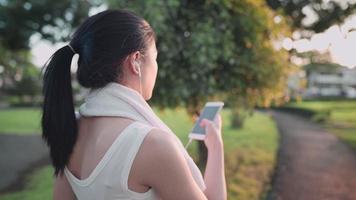 por cima do ombro atirou em mulher asiática usando smartphone enquanto caminhava pela trilha no jardim ao ar livre durante a hora do sol quente, relaxando após o treino, limpando o suor do rosto, estilo de vida de tecnologia video