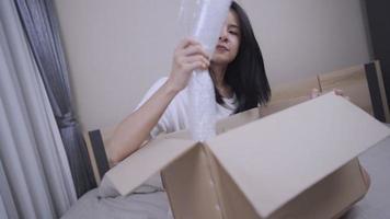 junge asiatische empfängerin setzt sich auf das bett, entpackt lieferpaket, online-einkaufspaket, lagerung, verwaltet den umzug, bleibt zu hause, öffnet paketbox, versandgebührensteuerservice video