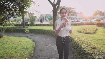 asian sporty woman using smartphone while walking down on the walking path at the recreation outdoor park during sunset hour, relaxing after workout, wiping sweat from face, new normal modern life video