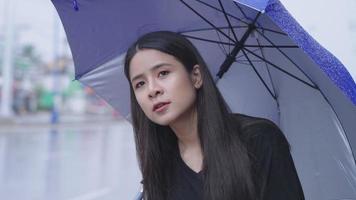 uma linda mulher do leste asiático fica sozinha na estrada esperando táxi ou ônibus com pressa chovendo tempo, segurando guarda-chuva na chuva, pressa hora do rush, mudança climática tropical, estilo urbano na estação chuvosa