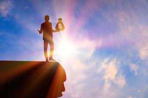 Silhouette businessmen standing on the stone looking distance and holding money bags with a dollar sign in their right hand with orange sunrise. photo