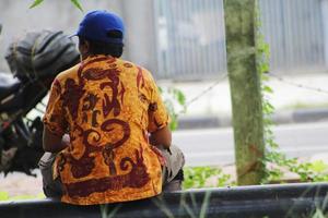 A middle-aged man is sitting on the side of the street in Jakarta, wearing Indonesian batik clothes