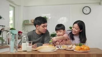 desayuno familiar asiático en casa. padres e hijos disfrutan comiendo juntos, conversando entre risas y buen ambiente. el padre juega con el hijo juguetonamente en la mesa de la cocina. video