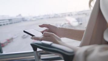 primo piano le mani della donna d'affari utilizzando lo smartphone che invia messaggi all'interno del gate di partenza del terminal dell'aeroporto durante l'attesa del volo, telefono cellulare che controlla le e-mail, viaggio di lavoro, cellulare e bagagli da viaggio video