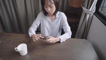 attraktiv asiatisk kvinna med kort hår som glider på smartphone med att läsa onlineinformation i matsalen, tillgång till internetkunskaper, kommunikation på avstånd, koppla av underhållande aktivitet video