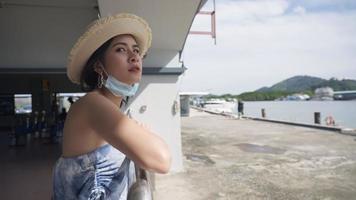 un hermoso joven turista asiático con ropa de sombrero de sol de moda y mascarilla médica sonriendo y mirando a la cámara mientras está feliz de pie solo al aire libre en una entrada, gente y un nuevo estilo de vida normal video