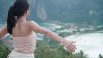 Rear view of young tourist woman enjoys fresh wind on tropical rainforest viewpoint, looks at green mountain cliff landscape, a fashionable summer wears, cliff observation activity, natural eco travel
