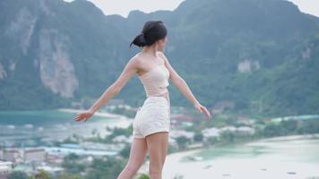 feliz, atractiva y confiada mujer asiática joven turista de caminata baila lentamente estirando los brazos mientras está de pie en el mirador de la isla en la cima de la colina, un viaje ecológico natural, escapa del concepto de ciudad