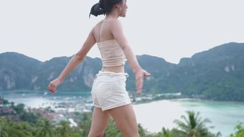 Following shot on cheerful asian model dancing around herself on a tropical sea coast viewpoint, active activist relaxing looks at green mountain cliff landscape, natural conservation tourism video