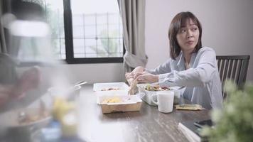 asiatisk arbetande kvinna som äter lunch hemma, leverans av matlåda för engångsbehållare, använder pinnar och sked, matleveransservice take away mat, nytt normalt hemdistansliv, håll avstånd video