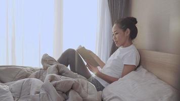 jovem mulher asiática confortavelmente inclinando o corpo na cama lendo o livro sozinho, atividade matinal produtiva, lençóis confortáveis do cobertor do quarto janela luz do dia, auto-educação, momento inspirador criativo
