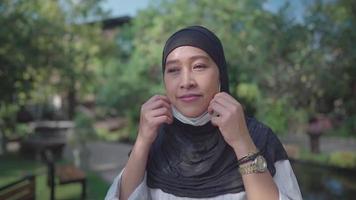 slow-motion shot van jonge aziatische moslimvrouw die een beschermend gezichtsmasker draagt. vrouw in hijab die op zonnige dag in het park staat, nieuwe normale coronavirus covid-19 pandemie, preventie van infectieziekten video