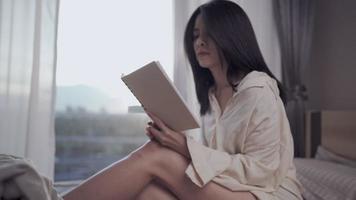 joven asiática sentada leyendo un libro sola en la cama, estudiando en casa pasando páginas, revisando el cuaderno de conferencias, el conocimiento de la lección, la luz del día en la ventana del dormitorio, la autoeducación, memorizando para el examen final video