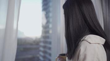 jovem mulher asiática tomando café da manhã depois de se levantar da cama, fêmea desfrutar de uma xícara de café e olhar pela janela em condomínio alto com vista urbana, relaxamento de manhã cedo despreocupado