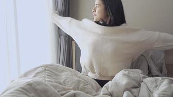 Young Asian woman getting up and Stretch arms on the bed, sunrise morning light, at home relaxing comfortable bedroom window curtains open, natural day light freshness, wearing sweater in warm winter
