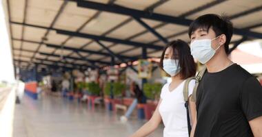 Side view of Young Asian traveler couple waiting the train at train station. Man and woman wearing protective masks, during Covid-19 emergency. Transportation, travel and social distancing concept. video