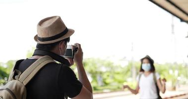 casal feliz viajante asiático com chapéu falando na câmera na estação de trem. jovem tirando fotos para a namorada. pessoas usando máscaras de proteção, durante a emergência covid-19. passatempo,