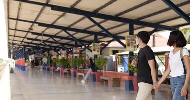 sidovy av unga asiatiska resenärspar som väntar på tåget vid tågstationen. man och kvinna som bär skyddsmasker under covid-19-nödsituationen. transport, resor och social distanseringskoncept. video