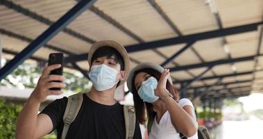 Happy asian traveler couple with hat video call on smartphone at train station. Young Man taking a pictures on mobile phone. People wearing protective masks, during Covid-19 emergency.