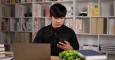 vista lateral del retrato de un hombre de negocios asiático que trabaja en una laptop y habla por teléfono en la oficina de su casa. joven autónomo escribiendo en el cuaderno de la computadora mientras está sentado en el escritorio en casa. concepto de distanciamiento social. video