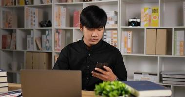 vista lateral del retrato de un hombre de negocios asiático que trabaja en una laptop y habla por teléfono en la oficina de su casa. joven autónomo escribiendo en el cuaderno de la computadora mientras está sentado en el escritorio en casa. concepto de distanciamiento social. video