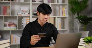 portrait d'un jeune homme asiatique a appuyé sur le code de la carte de crédit pour payer en ligne via un ordinateur portable au bureau dans la chambre. homme tenant une carte de crédit et faisant des achats en ligne. achats en ligne et concept de commerce électronique.