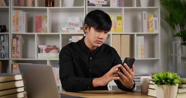 Young Asian businessman relaxing using phone on the table modern office. Bored manager surfing in internet. Tired male using cellphone at home office. Man browsing with mobile phone. video