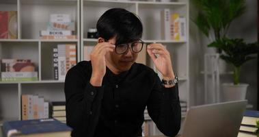 Tired young asian businessman eyeglasses stretching neck muscles while sitting at desk. Man doing neck massage feeling tired after work computer work sit at desk. Health care concept. video